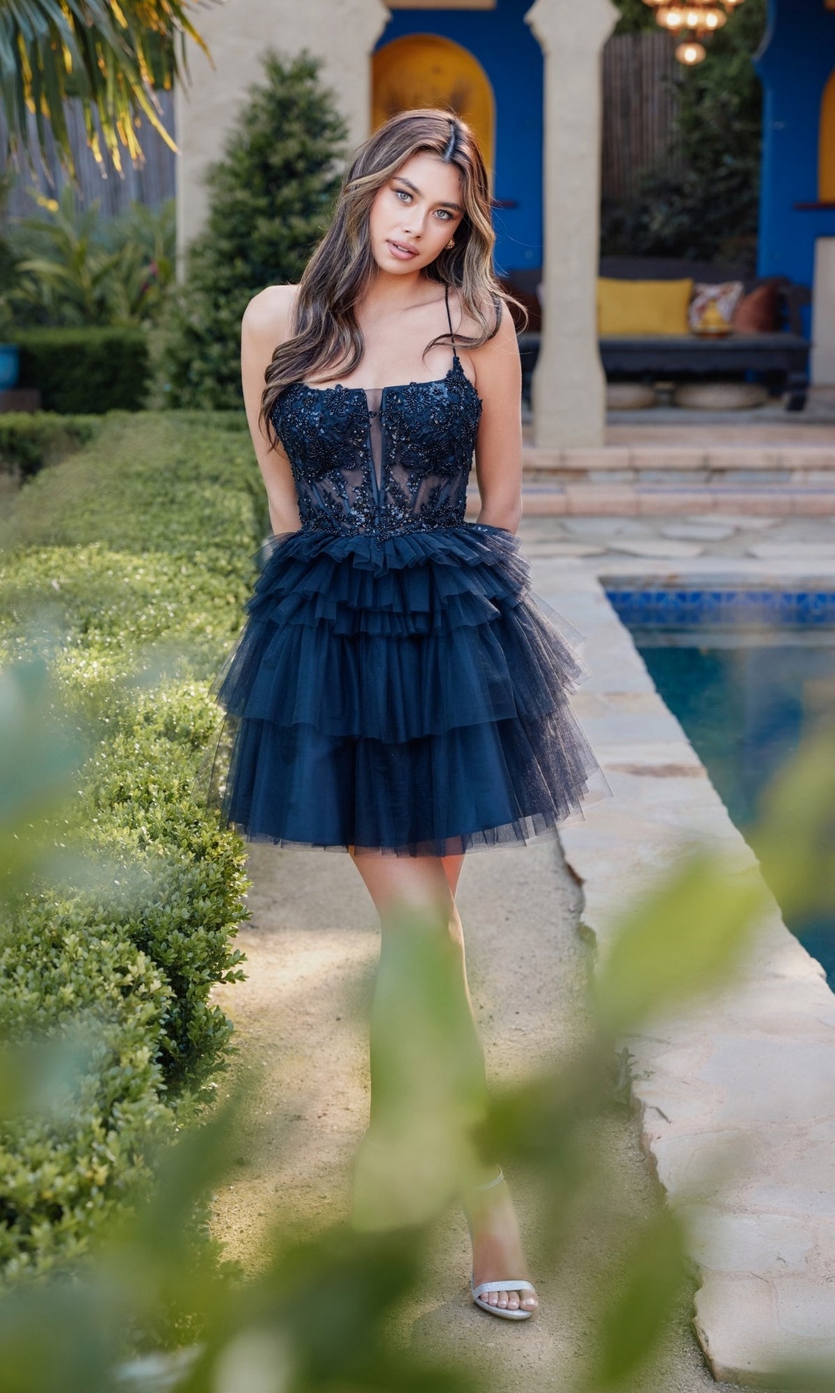 A woman wearing the Juliet JT935K short ruffled homecoming dress with a lace-up back stands on a garden path by a pool, surrounded by greenery and a building.